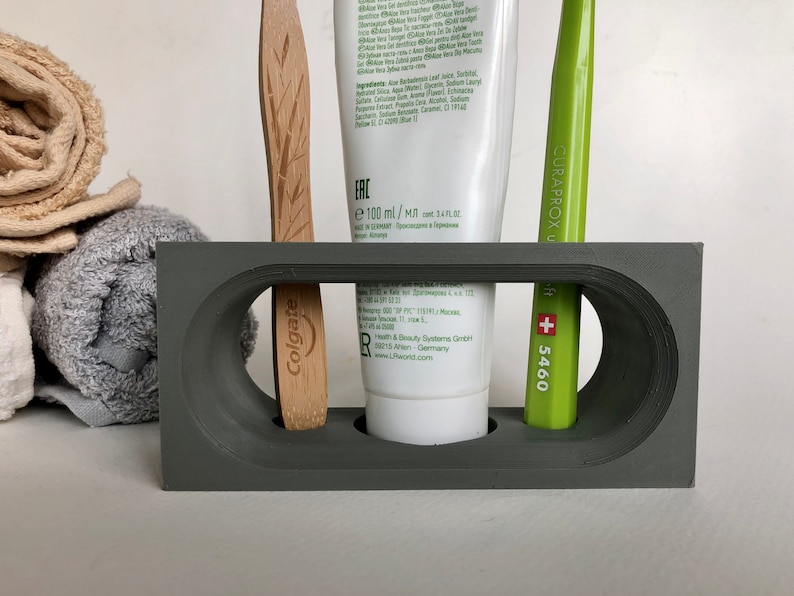 May include: Gray bathroom countertop organizer with three slots. A white tube of toothpaste with green lettering that says "ERC e 100 ml/Ml" is in the center slot. A green toothbrush with white lettering that says "CURAPROX +5460" is in the right slot. A wooden toothbrush with green lettering that says "Colgate" is in the left slot.