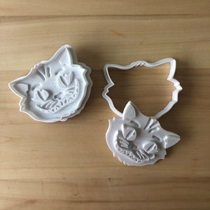 May include: Three white plastic cookie cutters in the shape of the Cheshire Cat's head. The cutters have detailed features, including the cat's grin, eyes, and striped fur. The cutters are on a light brown wooden surface.
