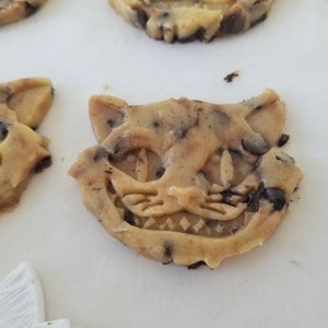 May include: Close-up of several chocolate chip cookies shaped like the Cheshire Cat from Alice in Wonderland. The cookies are light brown with dark chocolate chips and detailed facial features.