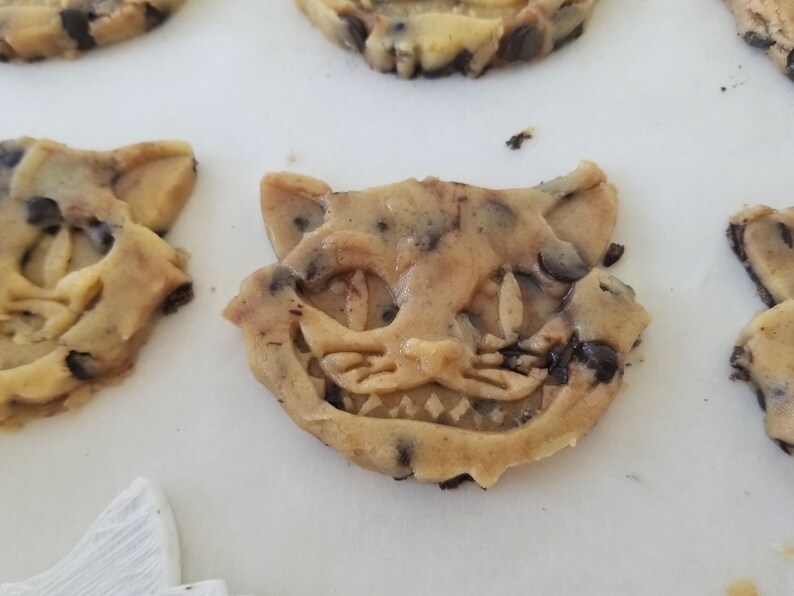 May include: Close-up of several chocolate chip cookies shaped like the Cheshire Cat from Alice in Wonderland. The cookies are light brown with dark chocolate chips and detailed facial features.