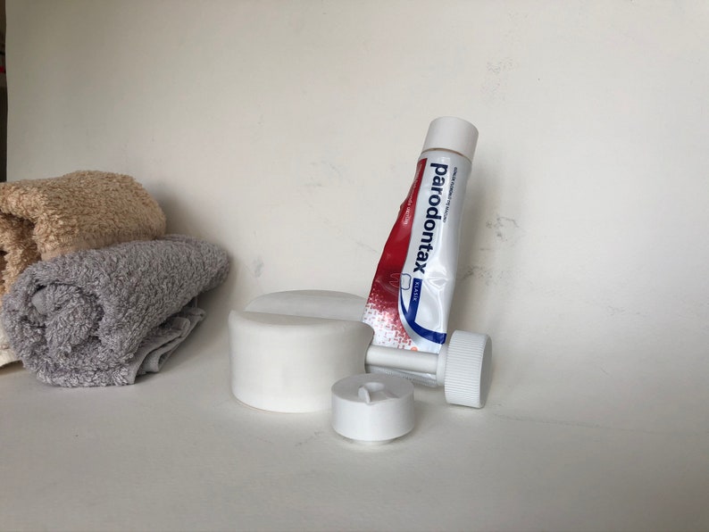 May include: A tube of Parodontax toothpaste with a red and white label, next to a white plastic toothpaste tube squeezer and two rolled towels. The toothpaste tube is partially squeezed. The towels are beige and gray.