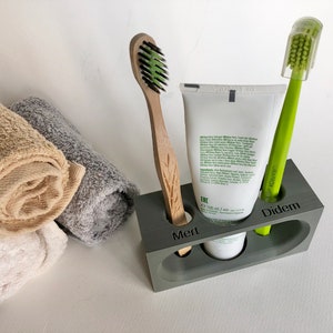 May include: A gray, rectangular toothbrush holder with three compartments. The holder has the names "Mert", "Diddem", and "Erc" engraved on the front. A white tube of toothpaste is in the middle compartment, a green toothbrush is in the right compartment, and a wooden toothbrush is in the left compartment.