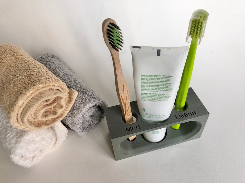 May include: A gray, rectangular toothbrush holder with three compartments. The holder has the names "Mert", "Diddem", and "Erc" engraved on the front. A white tube of toothpaste is in the middle compartment, a green toothbrush is in the right compartment, and a wooden toothbrush is in the left compartment.
