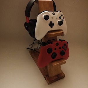 May include: A wooden stand with a headset, a white and a red video game controller. The stand is made of natural wood and has a simple design.