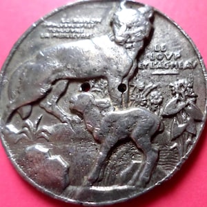 May include: A silver-toned metal medallion featuring a detailed relief of a large cat-like animal and a smaller lamb-like animal. The medallion has text around the edge and two small holes. The background is a pinkish-red color.