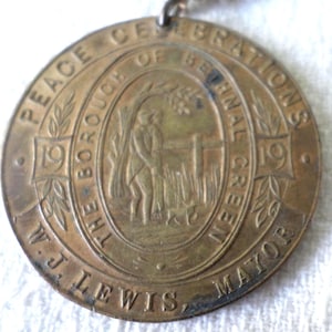 May include: A bronze-colored medal with the text "Peace Celebrations of Bethnal Green" and "1919" on the outer ring. The center features an illustration of a person and a dog near a fence. The text "W.J. Lewis, Mayor" is also visible.