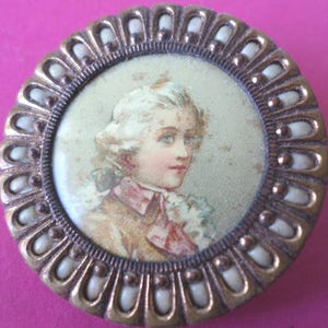 May include: A round, gold-toned button with a white porcelain rim. The button features a portrait of a young person with blonde hair, wearing a white collar and a brown jacket. The portrait is surrounded by a decorative border of white porcelain beads.
