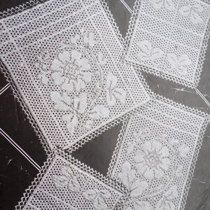 May include: Close-up of four white crocheted doilies with floral and leaf patterns. Each doily has a square shape with a decorative, scalloped edge. The doilies are arranged on a dark background.