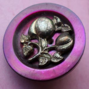 May include: A circular button featuring a silver fruit and leaf motif on a dark purple base. The button has an iridescent pink outer rim. Suitable for sewing or craft projects, this button has a decorative design.