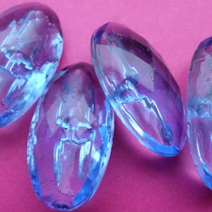 Peut inclure: Quatre perles de verre bleues et violettes, de forme ovale et facettée. Les perles sont transparentes, avec une teinte bleu clair sur les bords et un violet plus profond au centre. Elles sont disposées sur un fond rose.