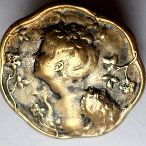 May include: Vintage brass button with an intricate relief design. It depicts a woman's profile, framed by floral motifs. The button's edge has a decorative, scalloped shape, enhancing its antique aesthetic. A classic piece.