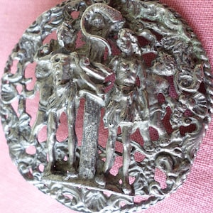 May include: A silver-toned metal brooch featuring a detailed design of three figures standing in front of a pillar. The figures are surrounded by intricate floral and scrollwork.