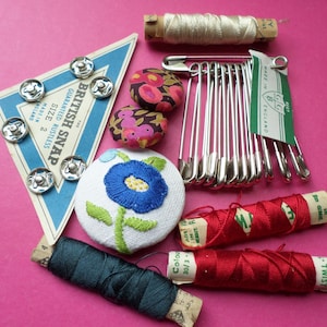 May include: A selection of haberdashery items on a pink surface. Features spools of thread, safety pins, fabric-covered buttons, an embroidered button, and a card of British Snap fasteners.