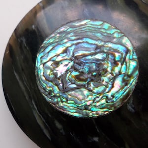 May include: Close-up of a round, iridescent abalone shell inlay set in a black, glossy surface. The abalone shell displays a vibrant mix of blues, greens, and purples, creating a shimmering effect. The black surface has a subtle, swirling pattern.