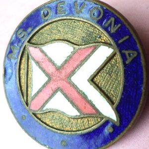 May include: Circular vintage pin with a blue enamel border and the text "M.S. DEVONIA". The center features a gold-colored textured background with a white and red cross design. The pin has a classic, aged appearance.