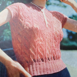 May include: A short-sleeved, coral-pink knit top featuring a vertical cable knit pattern and an intricate lace-like texture. The garment has a ribbed crew neckline and a wide, ribbed hemline that sits at the waist, providing a fitted silhouette.
