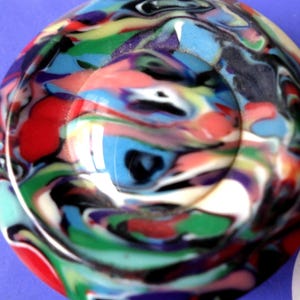 May include: A round, colorful object with a swirling pattern of red, blue, green, yellow, and black. The object has a glossy finish and a slight indentation in the center. The design resembles a marbled effect.
