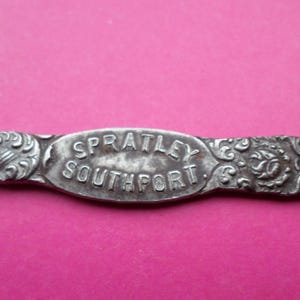 May include: A close-up of a silver metal object, featuring the text "SPRATLEY SOUTHPORT" in an oval. The object has decorative floral and shell patterns on either side of the text, set against a pink background.