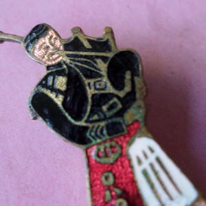 May include: A vintage metal pin depicting a figure in black clothing with gold detailing, a red coat, and a white sleeve. The pin has an intricate design and a small, horn-like extension. The figure seems to be playing a musical instrument.