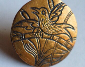 May include: A circular brass-toned button featuring an embossed design of a bird perched on a branch. The bird is depicted with wings slightly raised, and the background includes stylized foliage. The metal surface has a dark, recessed patina.