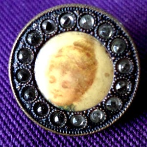 May include: A vintage button with a portrait in the center, surrounded by a ring of clear and dark gray stones. The button has a bronze-colored metal frame and is set against a purple background.