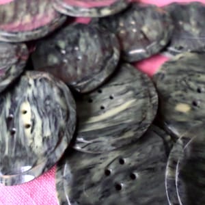 May include: A collection of large, round buttons with a marbled appearance. The buttons are primarily shades of gray and green, with a swirling pattern. Each button has two or four holes for sewing. The buttons are made of a hard, smooth material.