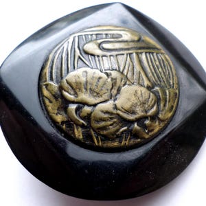 Peut inclure: Bouton noir en forme de losange avec un centre circulaire de couleur bronze. Le centre présente un relief détaillé de plantes aquatiques et de feuillage. La surface brillante et le design complexe suggèrent une esthétique vintage ou antique.