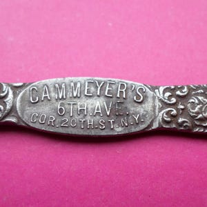 May include: Close-up of a silver metal object with the text "CAMMEYER'S 6TH AVE COR. 20TH ST. N.Y." in a rectangular center. The object has ornate floral and scrollwork designs on the ends.