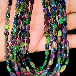 May include: A multi-strand necklace composed of numerous small, oval-shaped black opal beads. Each bead displays a dark base color with vibrant, iridescent flashes of green, red, blue, and yellow light throughout the polished stone surfaces.