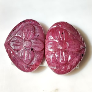 May include: Two carved ruby gemstones in the shape of a flower. The gemstones are a deep pink color and have a shiny, polished finish.
