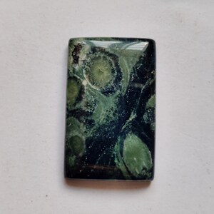 Könnte beinhalten: Ein grüner und schwarzer Edelstein-Cabochon mit einem einzigartigen, wirbelnden Muster. Der Stein ist rechteckig und hat eine polierte Oberfläche.