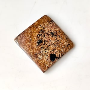 Puede incluir: Una piedra de forma cuadrada con una superficie pulida. La piedra presenta una mezcla de colores marrón, beige y negro, con un patrón moteado. Los bordes son ligeramente redondeados. La piedra está sobre un fondo blanco.