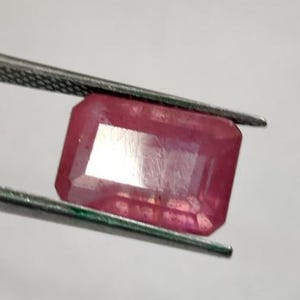 Może przedstawiać: Fasetowany, prostokątny kamień szlachetny rubinu trzymany pęsetą. Rubin ma żywy, różowo-czerwony kolor i odblaskową powierzchnię. Kamień szlachetny jest umieszczony na gładkim, białym tle.