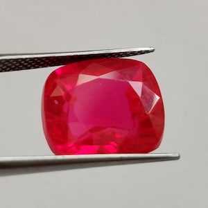 May include: A vibrant, cushion-cut ruby gemstone with a deep red hue and multifaceted surface, held securely between the tips of a pair of stainless steel metal tweezers. The gemstone displays a brilliant clarity and a polished, reflective finish.