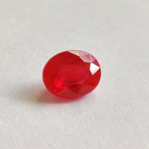 May include: A vibrant, oval-cut, red gemstone. The gem is displayed against a white backdrop, highlighting its colour and clarity. The facets of the stone create multiple reflective surfaces, enhancing its brilliance.