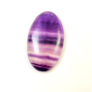 May include: An oval-shaped, polished gemstone with a layered appearance. The stone features bands of purple, lavender, and white hues. The gemstone is set against a white background, highlighting its color variations and smooth texture.
