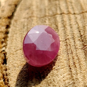 May include: A pink oval-shaped gemstone with a faceted cut. The gemstone is set against a light brown wooden background.