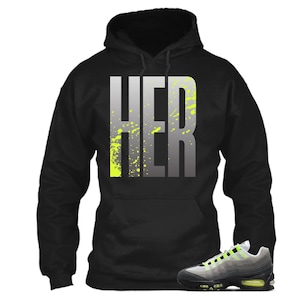 Puede incluir: Sudadera con capucha negra con la palabra "HER" en letras grises grandes con salpicaduras de color amarillo ne&oacute;n. Un par de zapatillas deportivas grises, negras y amarillo ne&oacute;n en primer plano.