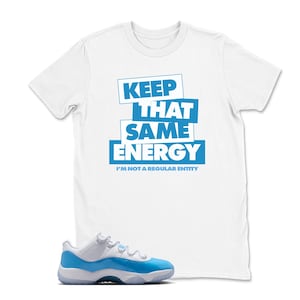 May include: White t-shirt with the text "KEEP THAT SAME ENERGY" in blue block letters. Below the text is the phrase "I'M NOT A REGULAR ENTITY". A pair of blue and white sneakers is in the foreground.