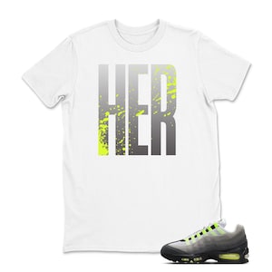 Puede incluir: Camiseta blanca con la palabra "HER" en degradado gris y salpicaduras verde ne&oacute;n. Un par de zapatillas deportivas grises, negras y verde ne&oacute;n est&aacute;n en primer plano, complementando el dise&ntilde;o de la camiseta.
