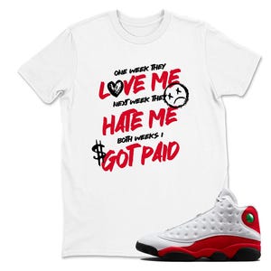 May include: White t-shirt with the text "ONE WEEK THEY LOVE ME, NEXT WEEK THEY HATE ME, BOTH WEEKS I GOT PAID" in red and black. A pair of white, red, and black sneakers is in the foreground.