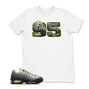 May include: White t-shirt with a graphic of the number "95" in a sneaker design, with black, gray, and neon green accents. A matching sneaker is in the foreground, featuring a black, gray, and neon green color scheme.