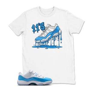 May include: White t-shirt featuring a cartoon illustration of a blue and white sneaker with the text "11's" in blue. A matching blue and white sneaker is in the foreground.