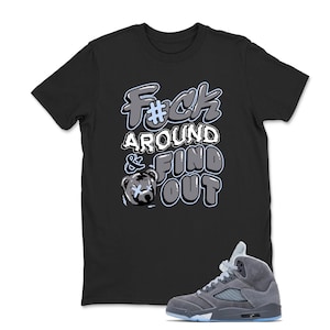May include: Black t-shirt with the text "F#ck Around & Find Out" in a bubble letter style. A cartoon bear with an X over its eyes is also featured. A gray and blue sneaker is in the bottom right corner.