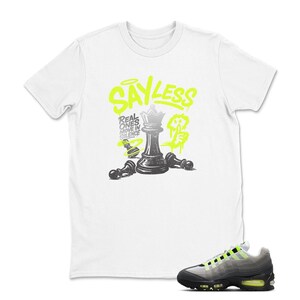 Puede incluir: Camiseta blanca con un gr&aacute;fico de ajedrez en negro y verde ne&oacute;n. El gr&aacute;fico incluye una pieza de ajedrez de reina y el texto "SAY LESS". Un par de zapatillas deportivas negras, grises y verde ne&oacute;n est&aacute;n en primer plano.