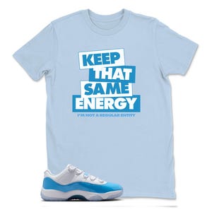 May include: A light blue t-shirt with the text "KEEP THAT SAME ENERGY" in white and blue block letters. Below the text is the phrase "I'M NOT A REGULAR ENTITY". A pair of blue and white sneakers is in the foreground.