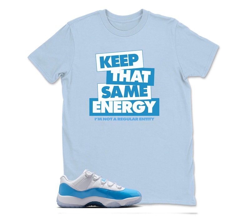 May include: A light blue t-shirt with the text "KEEP THAT SAME ENERGY" in white and blue block letters. Below the text is the phrase "I'M NOT A REGULAR ENTITY". A pair of blue and white sneakers is in the foreground.