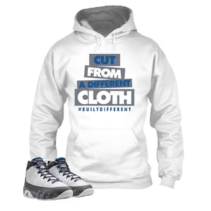 May include: White hoodie with the text "CUT FROM A DIFFERENT CLOTH" in blue and gray. The hoodie is paired with black, white, and blue sneakers. The phrase "#BUILTDIFFERENT" is below the main text.