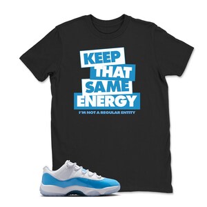 May include: Black t-shirt with the text "KEEP THAT SAME ENERGY" in white and blue. Below the text is "I'M NOT A REGULAR ENTITY". A pair of white and blue sneakers is in the foreground.
