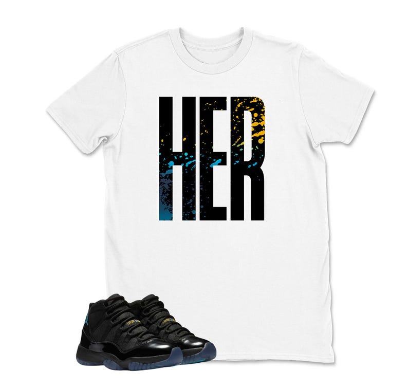 Op de afbeelding: Een wit t-shirt met het woord "HER" in grote zwarte blokletters, geaccentueerd met blauwe en gele verfspatten. Daaronder een paar zwarte en teal sneakers.